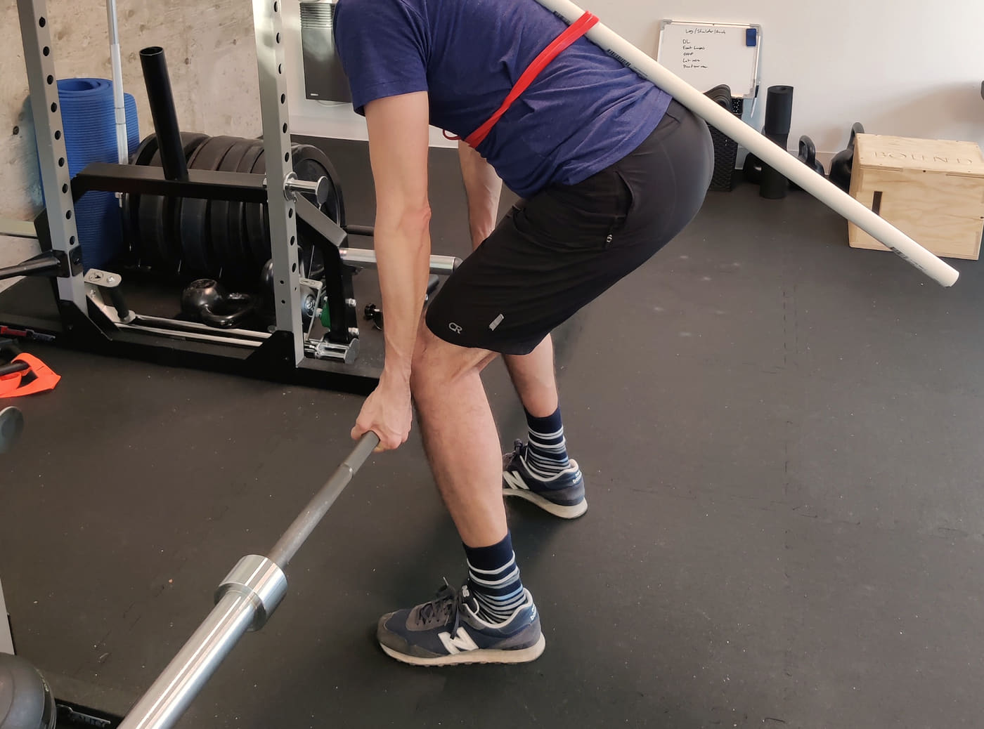 A hip hinge coached with a dowel along the spine and a barbell on the floor, next to the rack, sled and the session written on the whiteboard